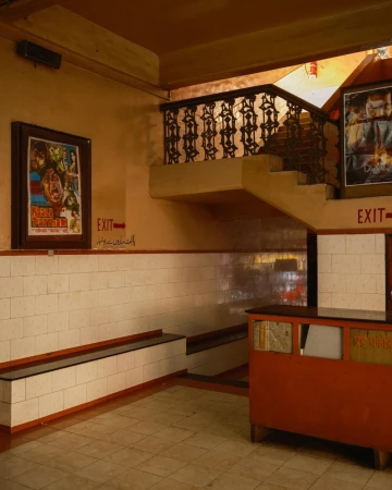 Indoor scene of a building lobby with a staircase, walls with white tiles, posters, and an exit sign with orange and beige color tones.