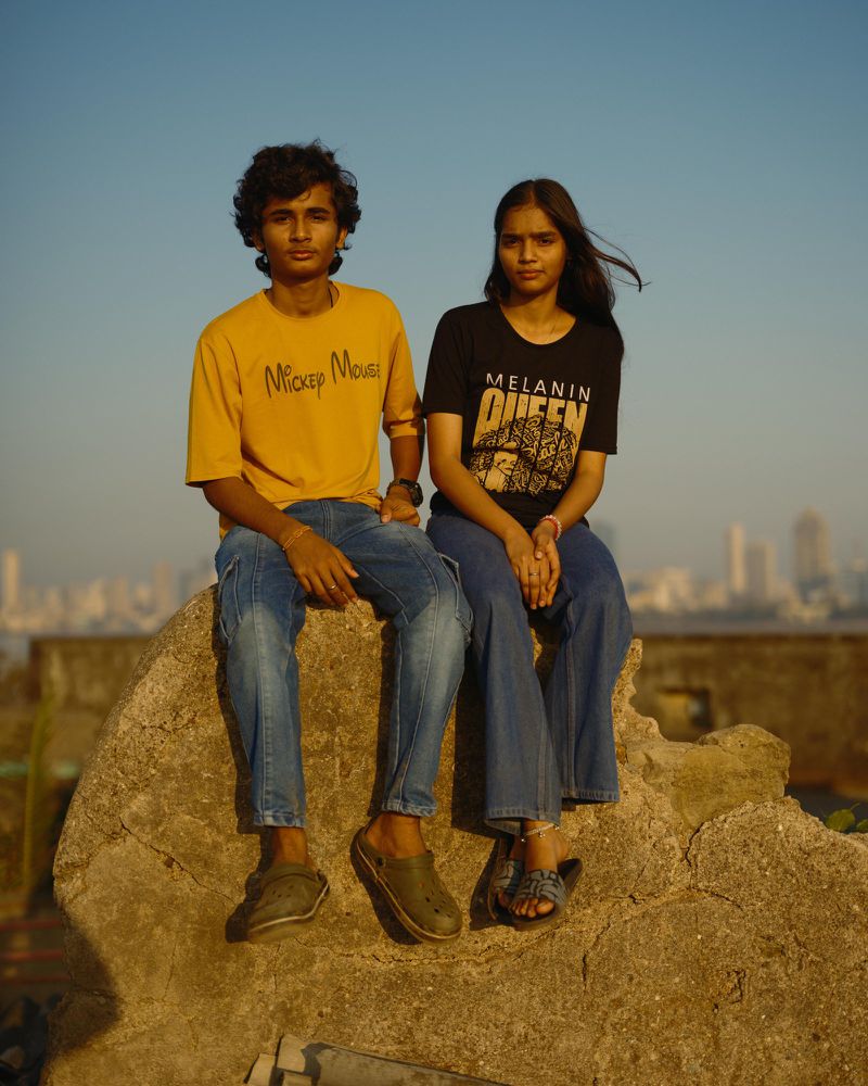 Two young adults, a male and female, are sitting on a rock formation with a cityscape in the background under a clear, bright sky.
