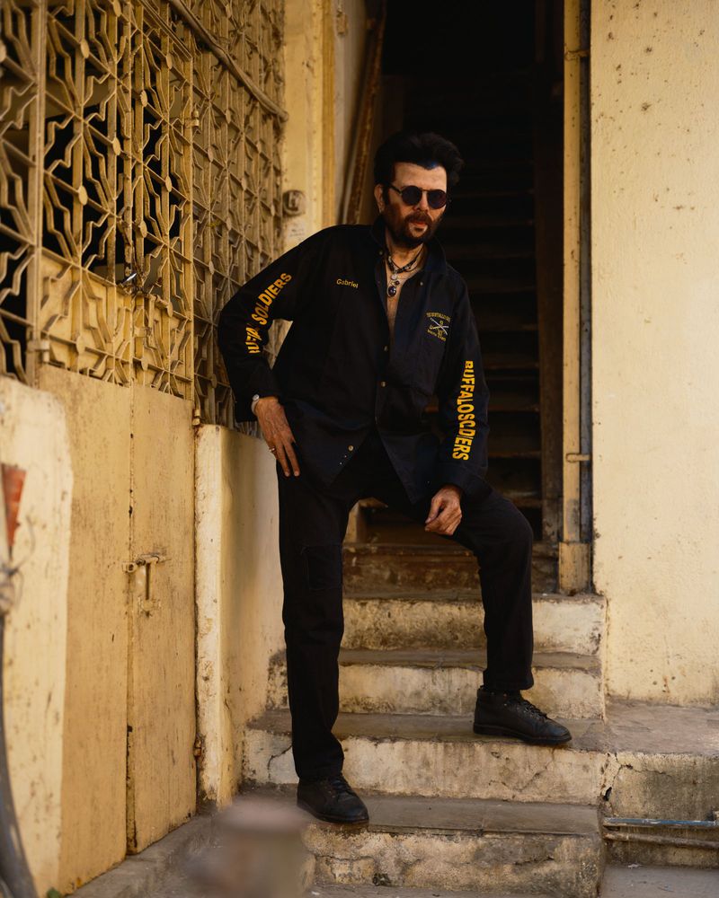 Man in black shirt and sunglasses poses on tan concrete stairs in daylight with patterned windows in the background in an outdoor shot.