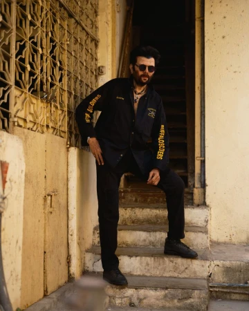 Man in black shirt and sunglasses poses on tan concrete stairs in daylight with patterned windows in the background in an outdoor shot.
