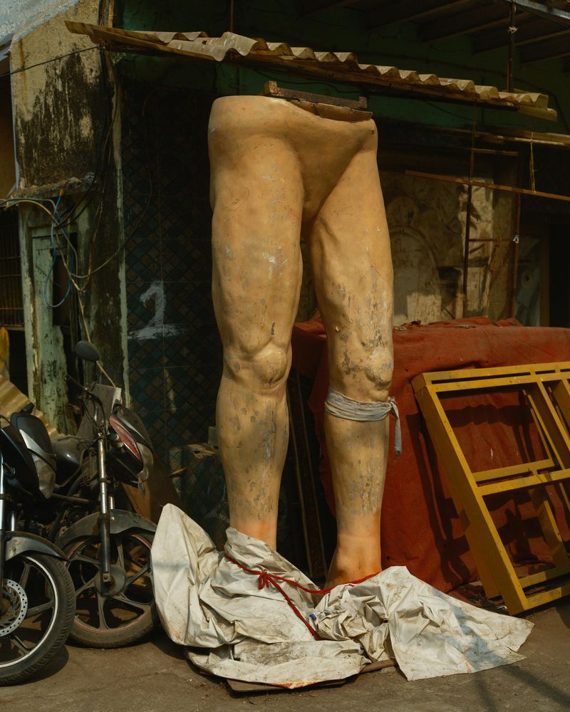 Outdoor shot of a weathered, headless statue of legs, propped up by a building, with motorcycles parked nearby, creating an urban scene.