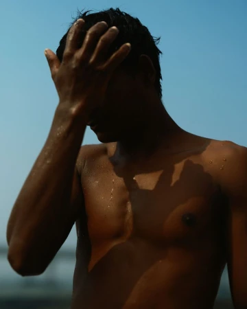 A shirtless man wipes his wet hair away from his face, illuminated by bright sunlight against a clear sky in an outdoor setting.
