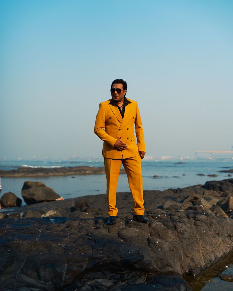 Adult man in a yellow suit standing on rock formation with ocean and sky in the background, fashion portrait on a sunny day.