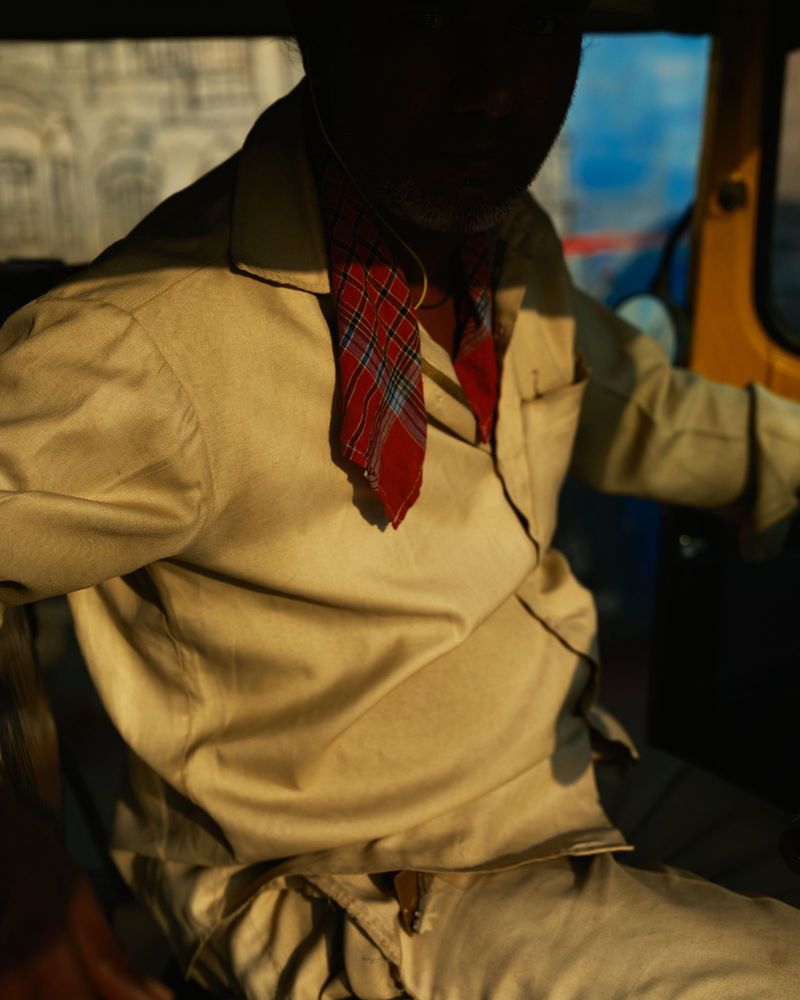 Man in beige shirt driving auto rickshaw. Red plaid scarf. The vehicle is yellow and the image is shot at eye-level, medium distance.