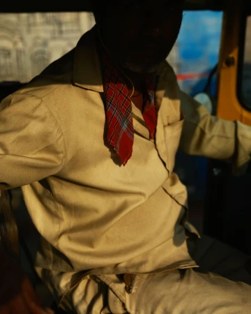 Man in beige shirt driving auto rickshaw. Red plaid scarf. The vehicle is yellow and the image is shot at eye-level, medium distance.