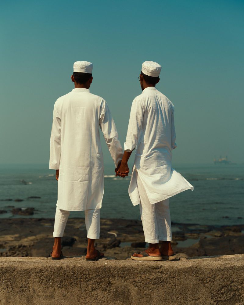 Two men in white clothing stand hand-in-hand against a sea backdrop on a sunny day, creating a serene and contemplative scene. 