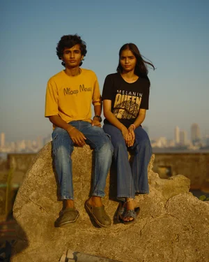 A young South Asian man in a yellow t-shirt and a young woman in a black t-shirt sitting together on a rock outdoors with a city skyline