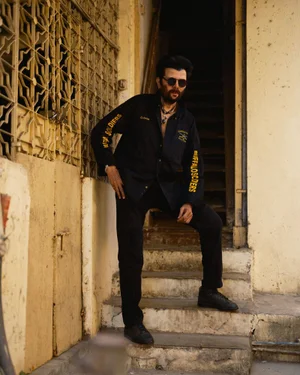 Man wearing sunglasses and a black long-sleeve shirt posing on outdoor concrete stairs beside a yellow patterned gate.