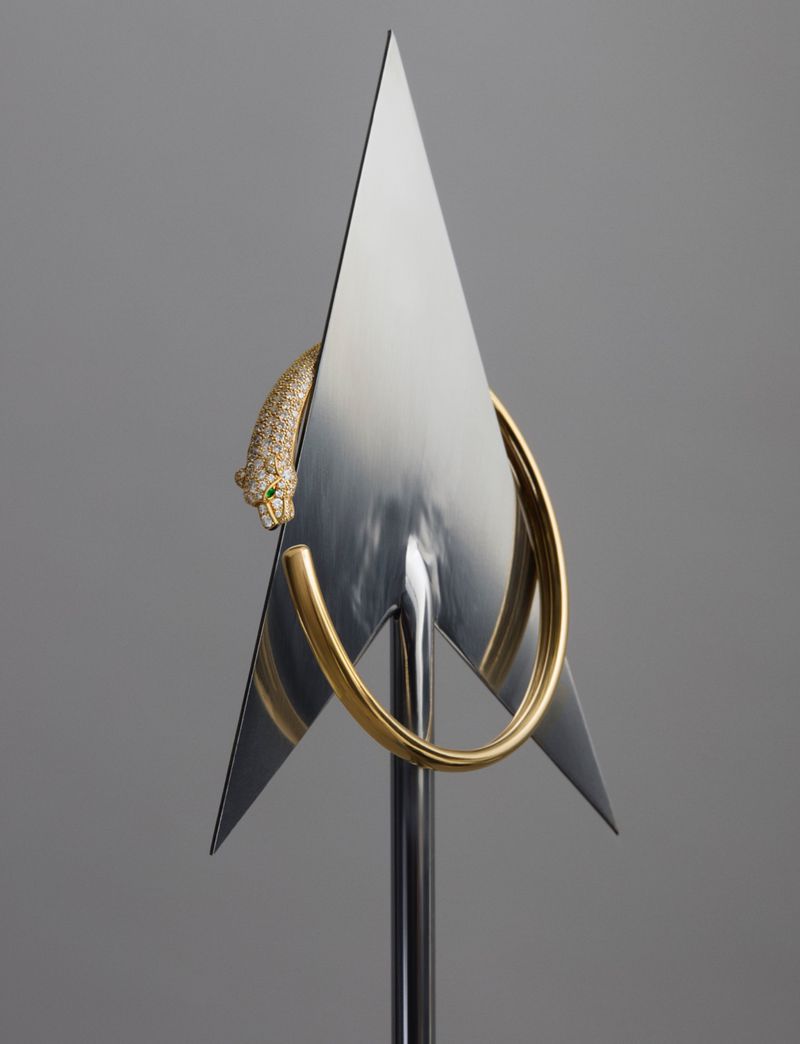 A golden panther sculpture with green eyes wraps around a metal hoop with a triangular metal design, standing on a sleek metal stand.