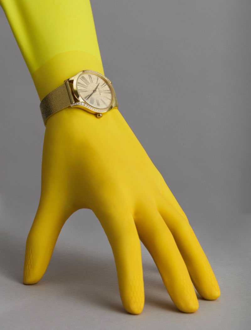 Wristwatch on gloved hand in studio setting. Gold watch with mesh strap on yellow gloved hand against a gray backdrop.