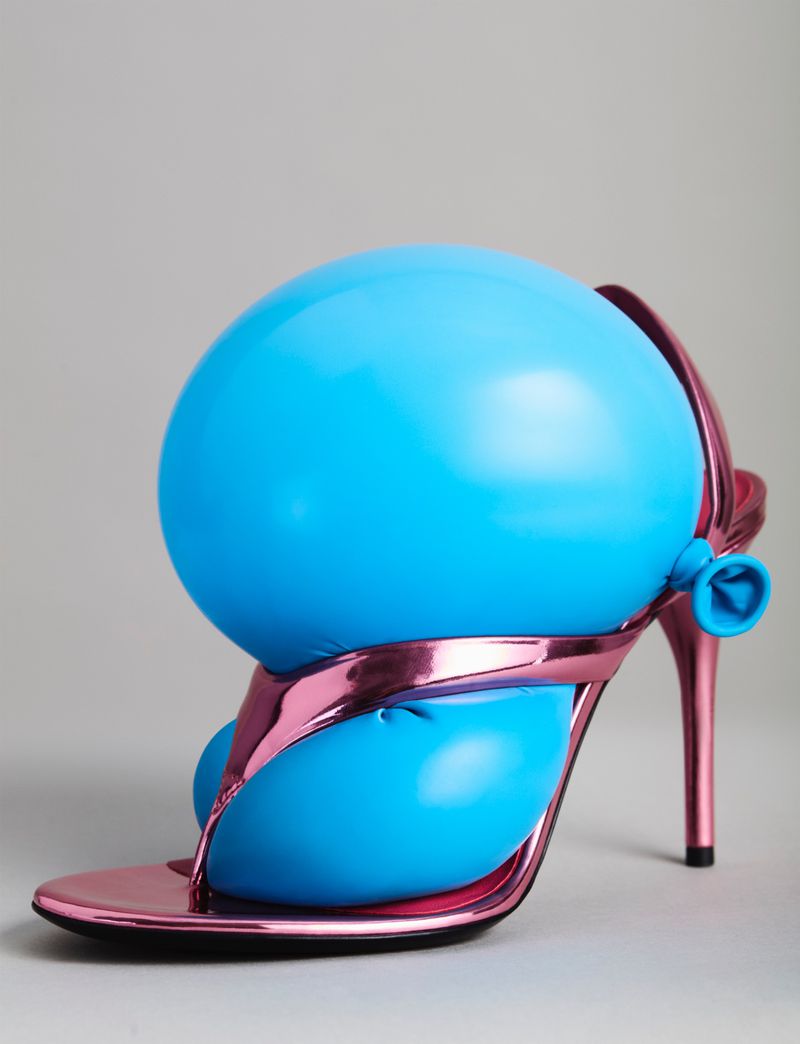 Stiletto heel shoe with pink metallic straps is filled and distorted with a large blue balloon in a studio setting.
