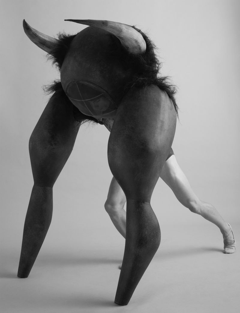 Black and white studio shot of a person wearing a dark bull-like costume with large horns, posed against a plain background.
