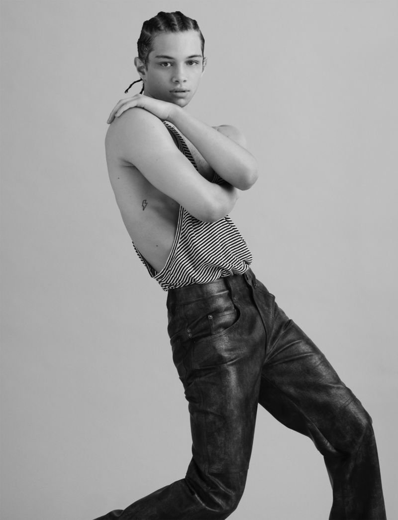 Black and white portrait of a young adult man with braided hair, striped shirt, and black leather pants against a grey studio background.