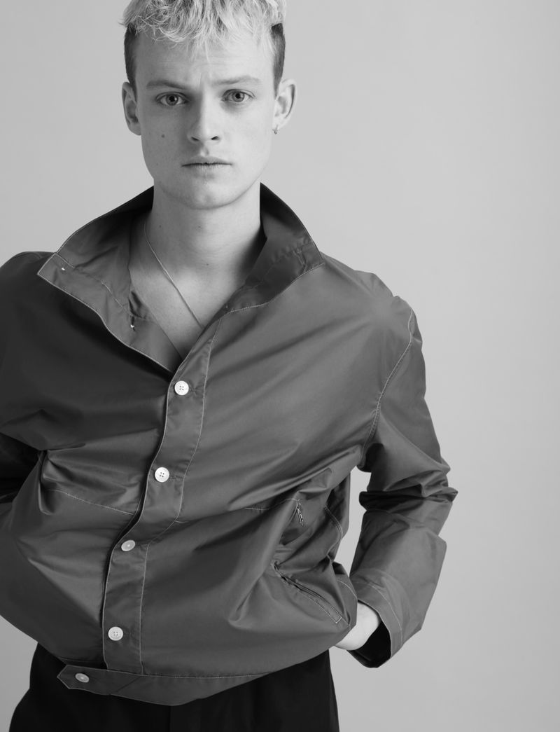 Portrait of a young adult male with fair skin and light blonde hair, wearing an open red button-down jacket and dark pants, in a studio setting.