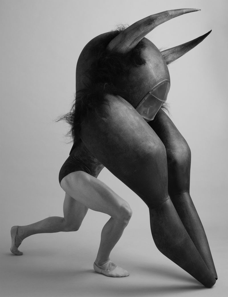 Black and white shot of a person in a leotard carrying a large, horned costume head, showcasing creativity and physical performance.
