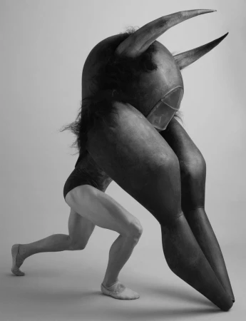 Black and white shot of a person in a leotard carrying a large, horned costume head, showcasing creativity and physical performance.