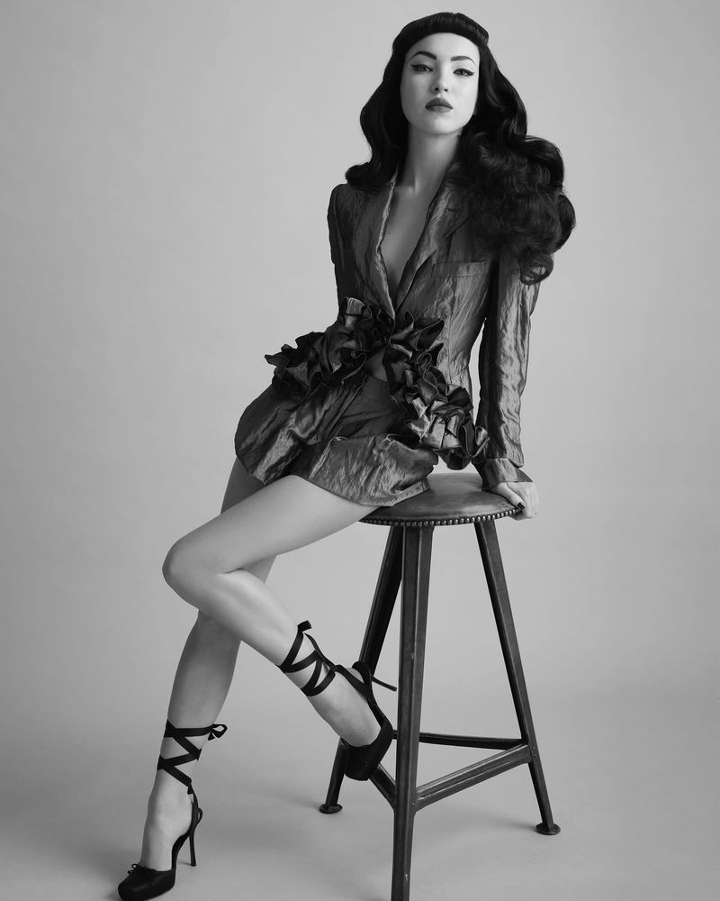 Black and white portrait of a stylish young woman with dark hair sitting on a stool wearing a jacket-dress and high heels with straps.