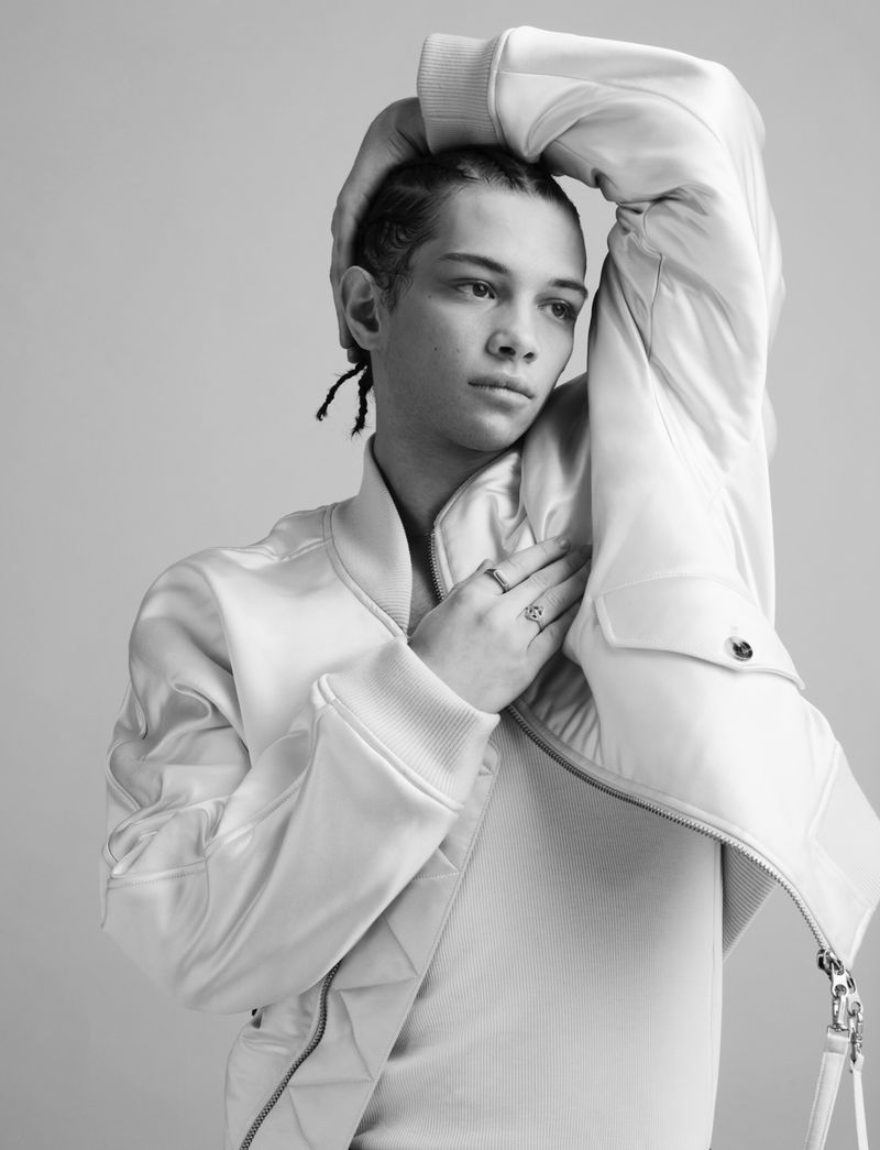 Studio portrait of a young adult male model in a light-colored jacket and top, with braids, posing against a neutral background.