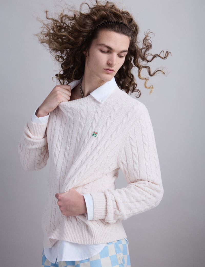 Portrait of a young adult male with long curly brown hair, wearing a cable knit sweater over a button-down shirt and checkered pants.