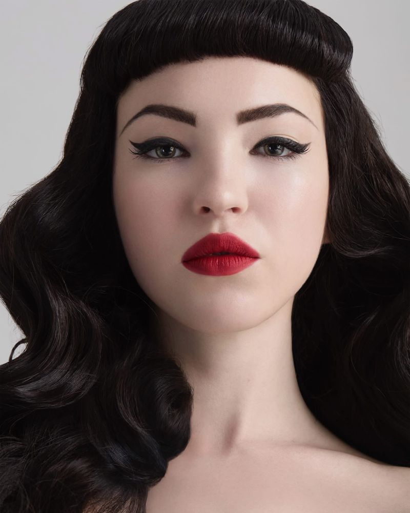 Close-up studio portrait of a woman with dark wavy hair, red lipstick, and winged eyeliner against a neutral background. Beauty shot.