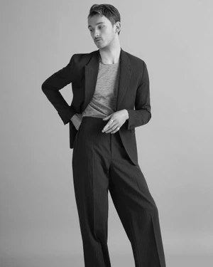 Black and white editorial photograph of a male model wearing a tailored pinstripe suit and grey t-shirt against a neutral background.