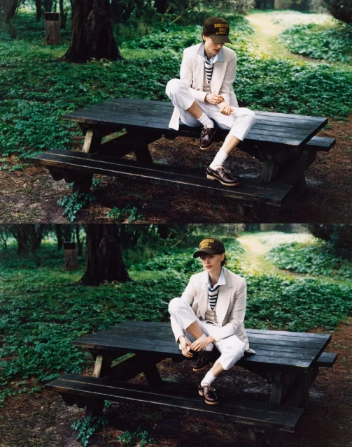 Woman in beige blazer, striped shirt, and baseball cap poses on a picnic table in a forest setting, capturing a relaxed and fashionable moment.