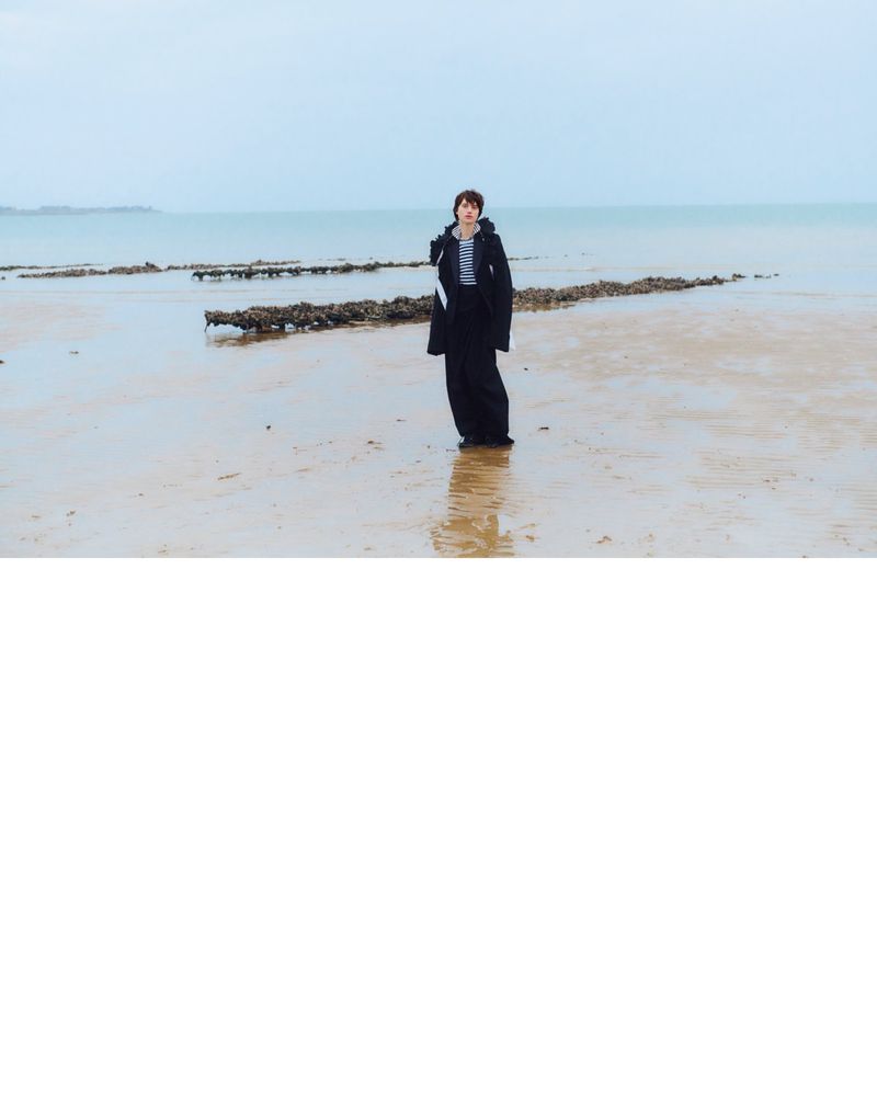 A person stands on a sandy beach with racks and ocean water under an overcast sky, wearing a dark coat and wide, dark pants.