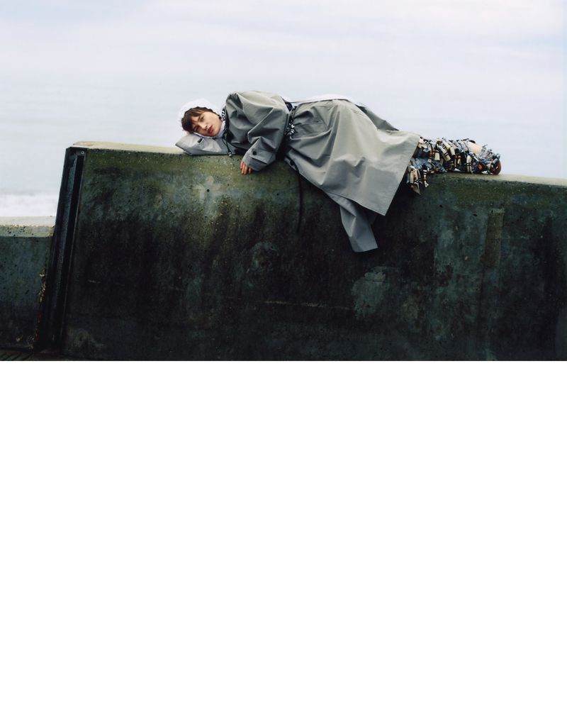 Adult female fashion model in grey coat lies on concrete wall near the ocean under cloudy sky in lifestyle editorial photography.