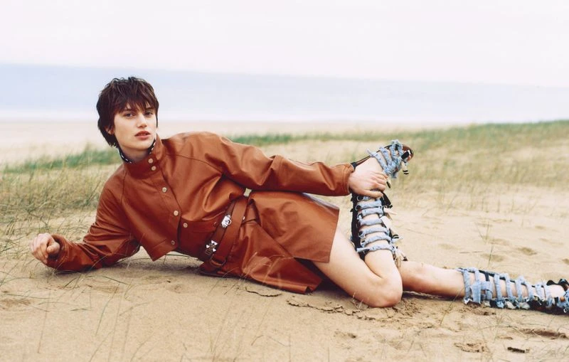 Fashion model in a rust-colored jacket and deconstructed denim leg coverings poses on a sandy beach near the ocean.