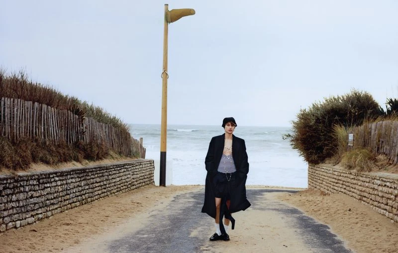 Fashion shot of a young man on a beach road, wearing a long black coat, shorts, and a striped shirt, posing near a vintage light post.