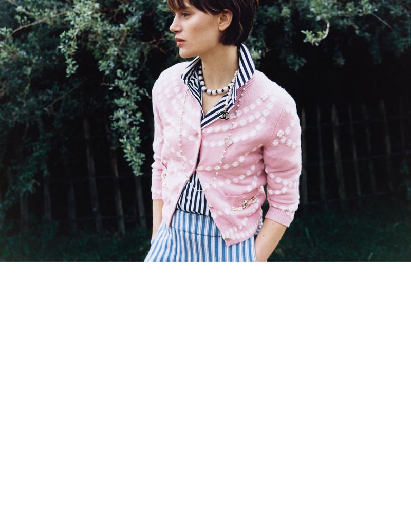Young woman in striped shirt and skirt wearing a light pink cardigan standing in a garden with green leafy bushes.