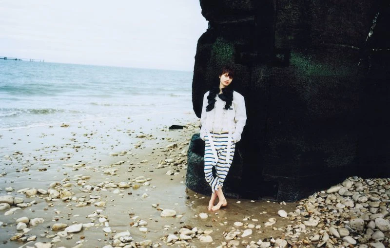 A young adult stands barefoot on a pebble beach near a black rock formation on an overcast day with the ocean in the background.