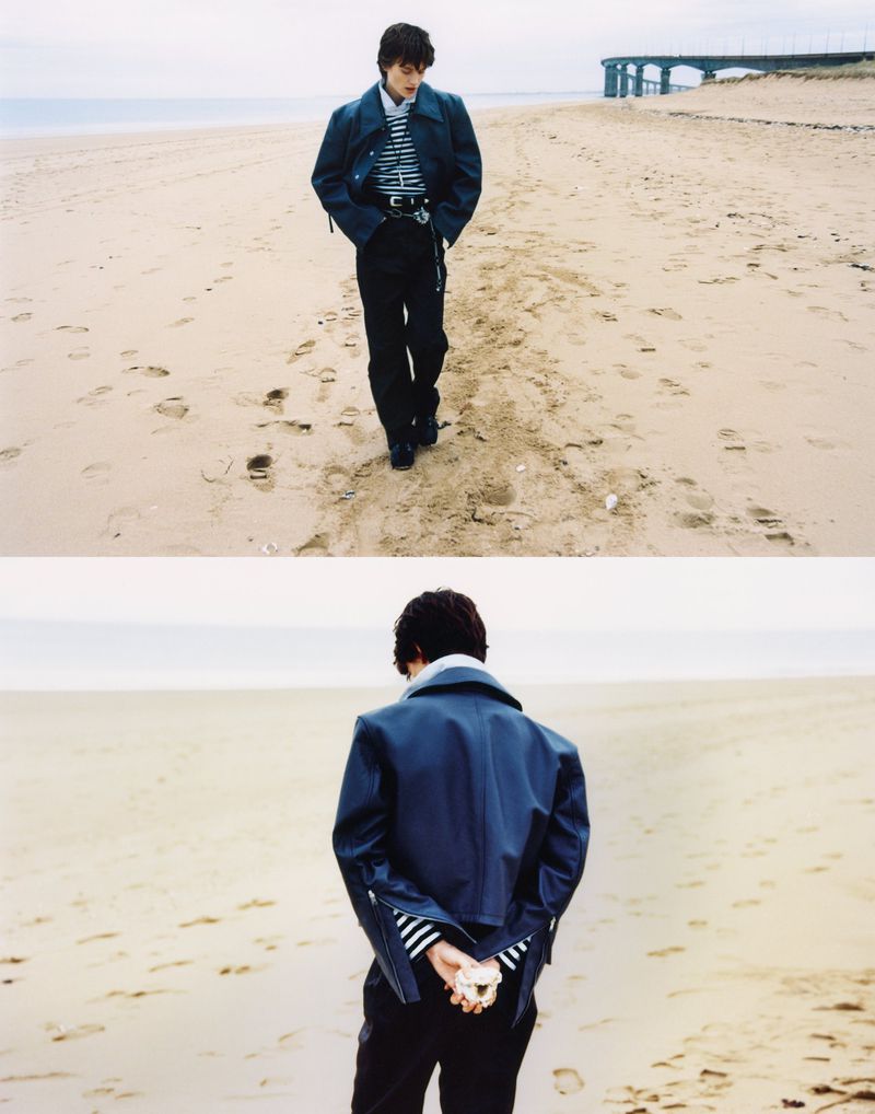 Person in jacket and striped shirt walking on a sandy beach near a pier, with footprints in the sand, under an overcast sky.