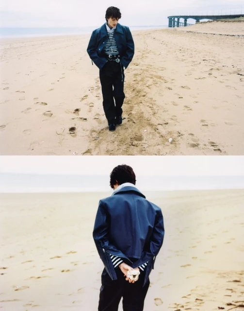 Person in jacket and striped shirt walking on a sandy beach near a pier, with footprints in the sand, under an overcast sky.