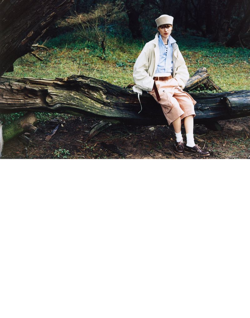 Young person sits on a fallen tree in a forest wearing vintage-style beige jacket, light blue shirt, salmon-colored shorts, white socks, and brown shoes.