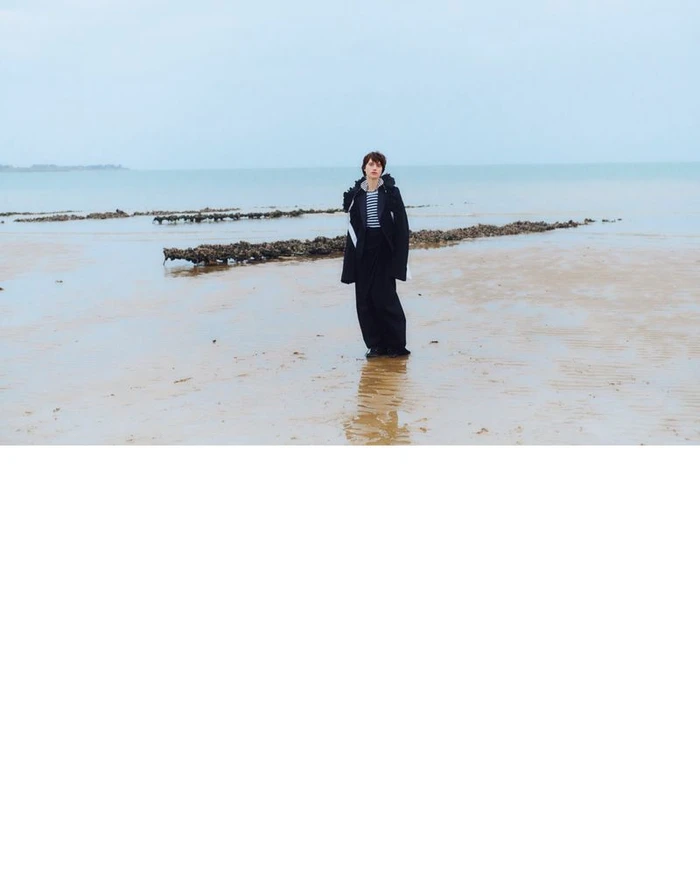 Model standing on a wet sandy beach wearing a dark coat and trousers under an overcast sky.