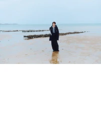 Model standing on a wet sandy beach wearing a dark coat and trousers under an overcast sky.