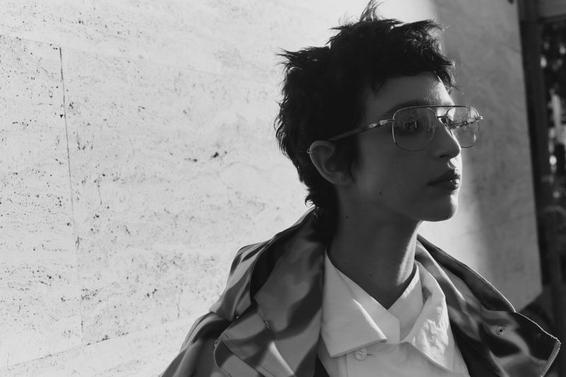 Black and white portrait of a person wearing glasses, a white button-down shirt, and a striped jacket, with a textured wall background.