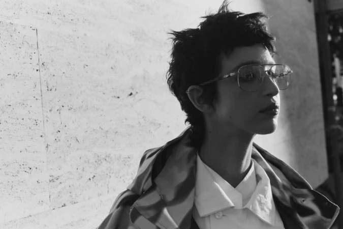 Black and white portrait of a person wearing glasses, a white button-down shirt, and a striped jacket, with a textured wall background.