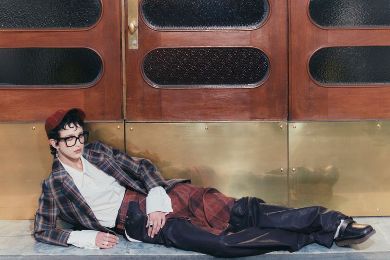 Stylish person in plaid blazer and trousers lying in front of a wooden and brass door, accessorized with glasses, cap, rings and earrings.