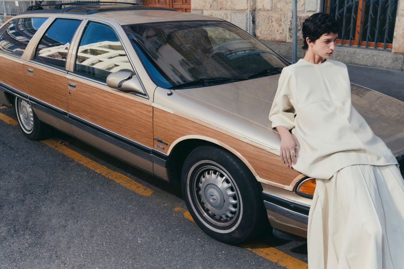 Woman in flowing dress leans on a wood-paneled station wagon against a city building backdrop, captured in soft, natural light.