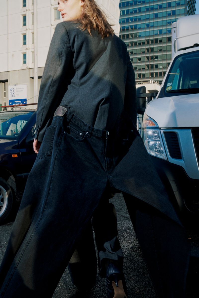 A woman poses in dark denim jacket and pants against a city street backdrop with parked cars and buildings in daytime lighting.