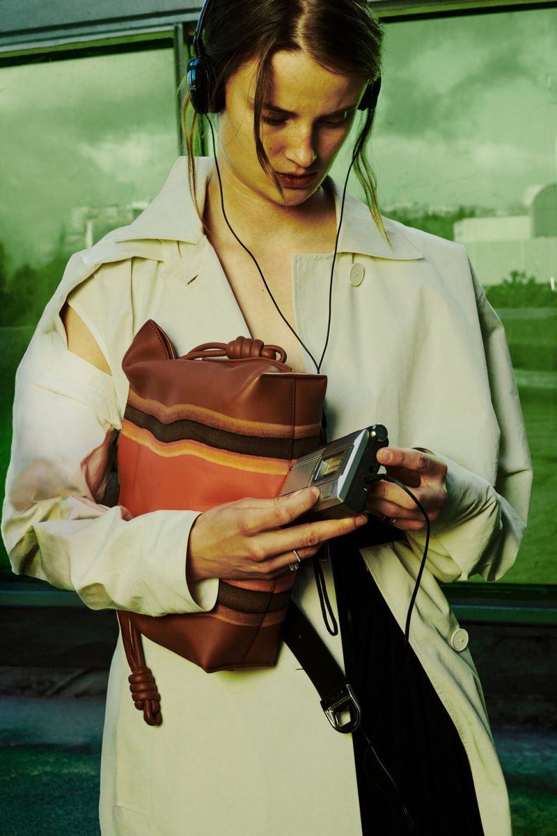 Woman in trench coat using a portable music player, wearing headphones, and carrying a brown leather backpack outdoors.