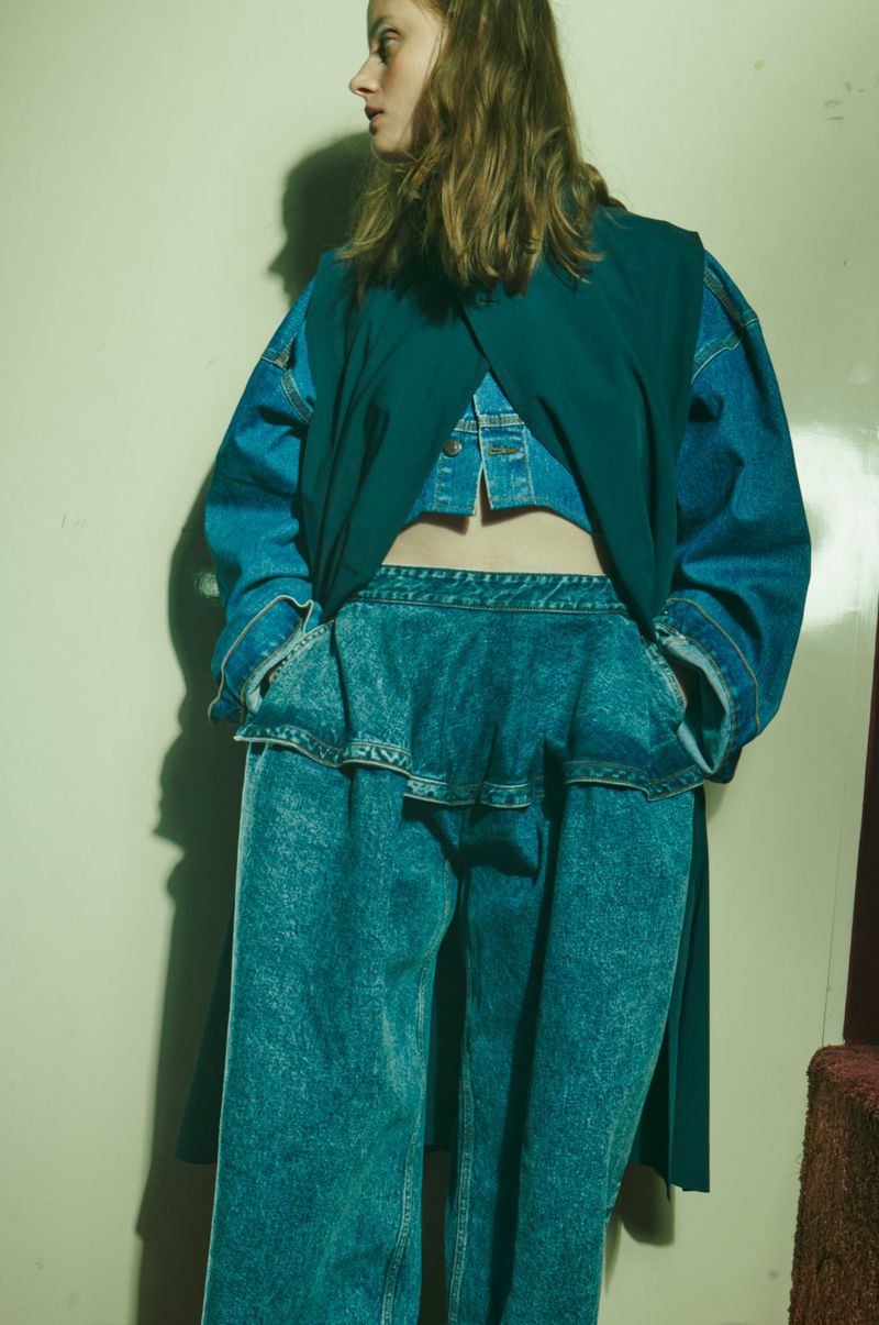 Fashionable woman in a striking teal top and blue jeans with pockets poses indoors against a cream wall, showcasing an edgy aesthetic.