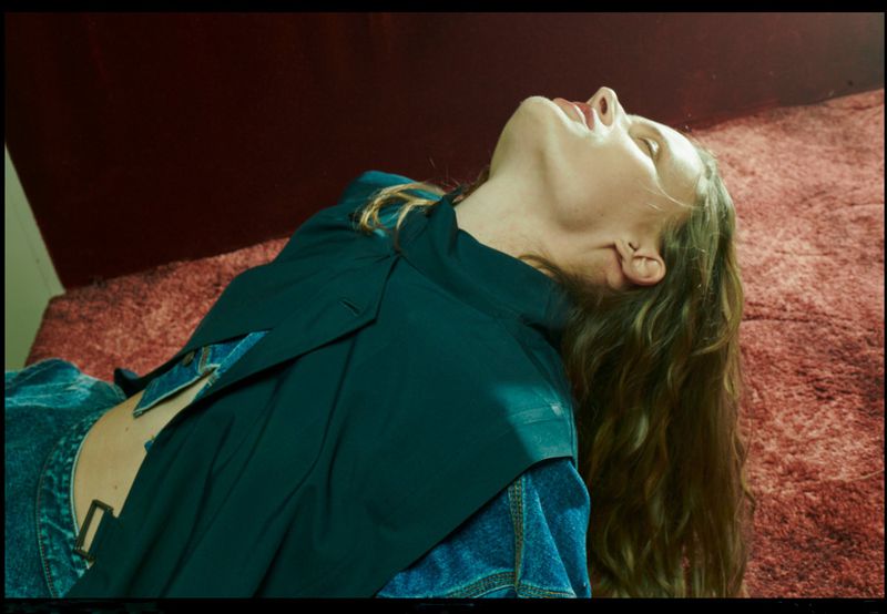 Adult female lying on a textured surface wearing a denim jacket and blazer with her head tilted back in a close-up shot.
