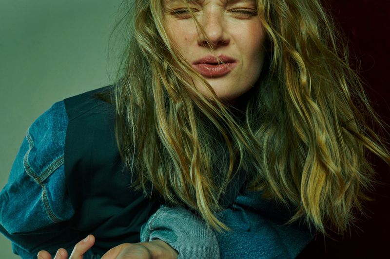 Close-up portrait of a blonde woman with a distressed expression wearing a denim jacket, posing in a studio with green and red lighting.