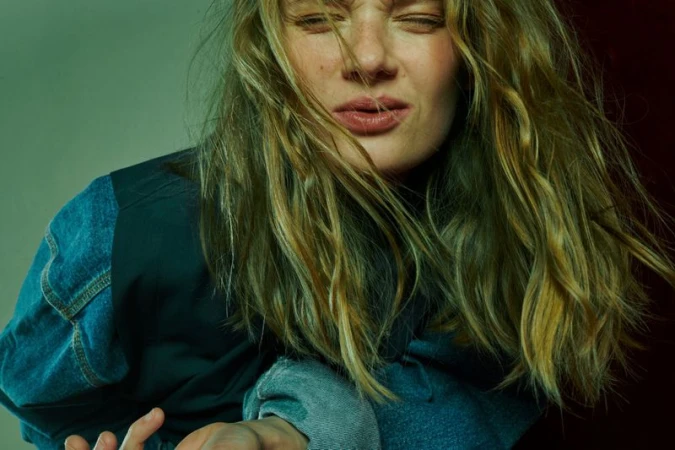 Close-up portrait of a blonde woman with a distressed expression wearing a denim jacket, posing in a studio with green and red lighting.