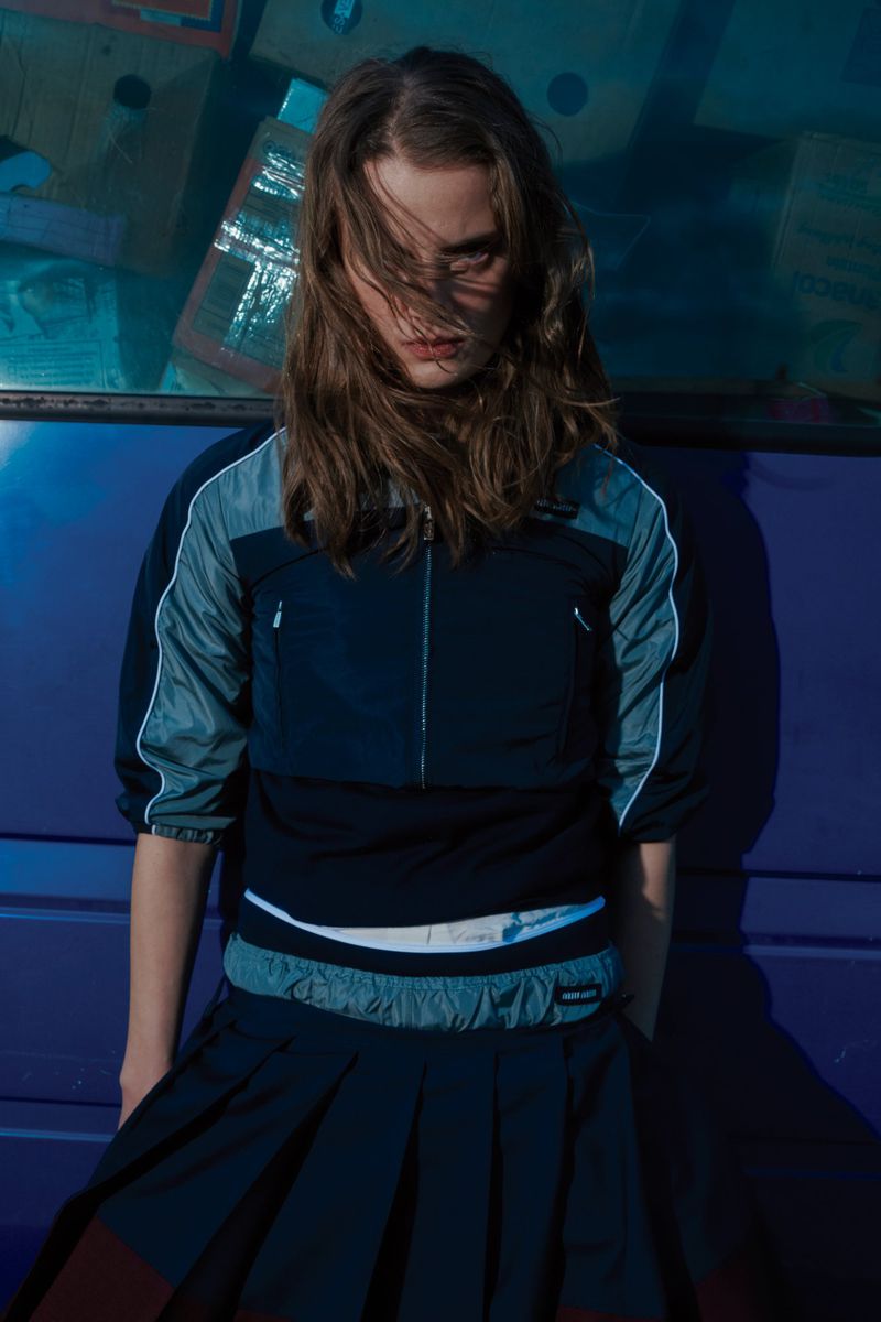 Teen girl in sporty top and pleated skirt poses against a purple wall, with diffused, natural light creating soft shadows and cool tones.