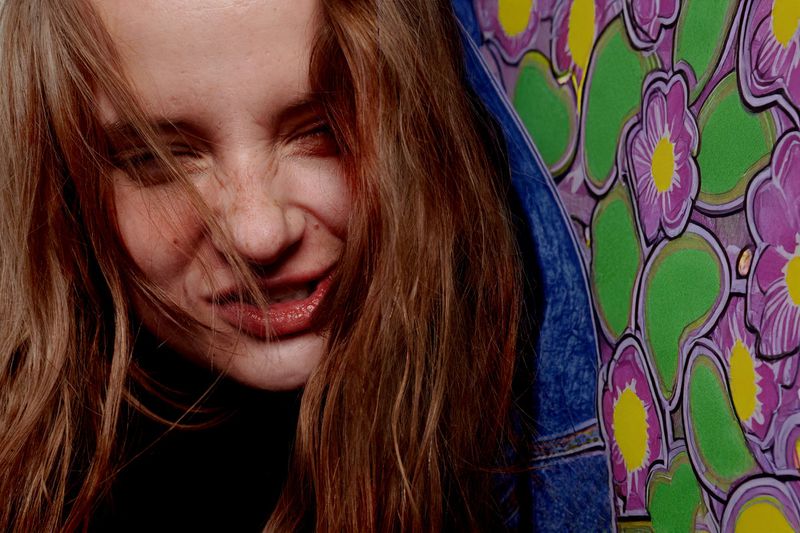 Close-up portrait of a young adult female with auburn hair and expressive facial expression, wearing a denim jacket near a floral painting.