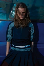 Model with long brown hair wearing a dark zip-up athletic top and pleated skirt against a blue vehicle background.